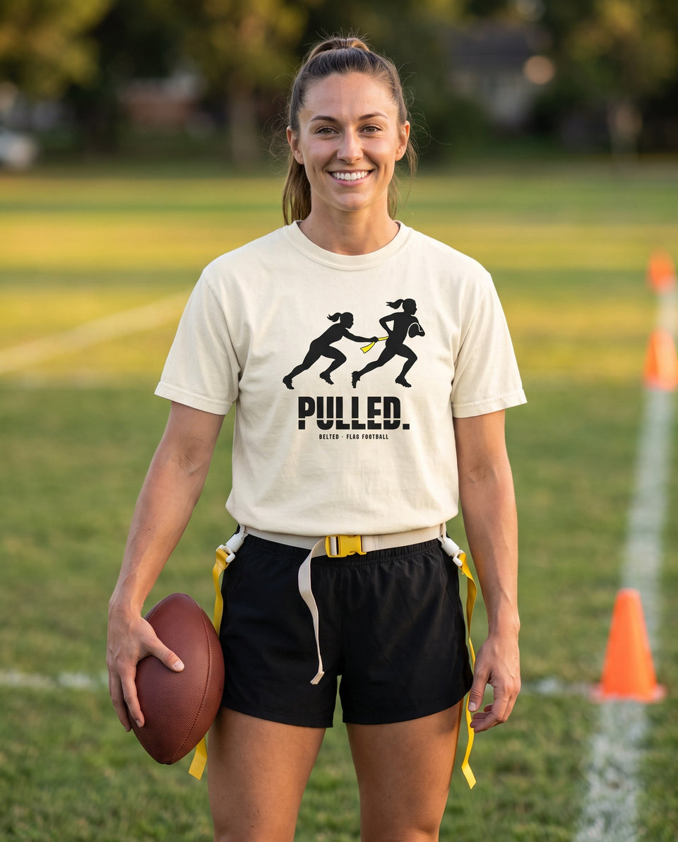 Ivory Pulled Tee on a smiling player holding a football, black two-player pull graphic, PULLED. wordmark, yellow flag