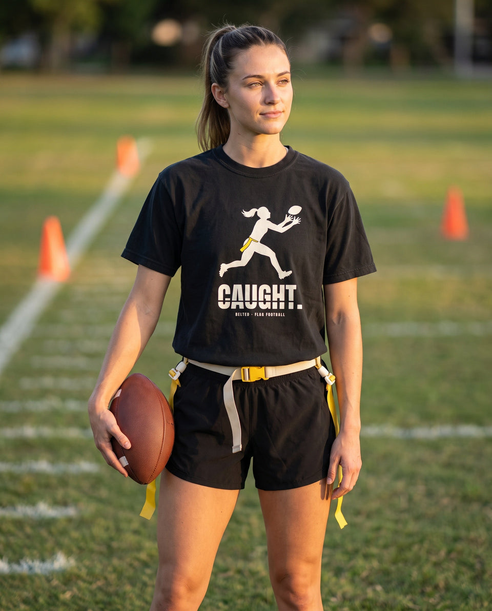 Caught Tee in black, worn by a flag football player holding a ball on a field, Ivory CAUGHT. print with receiver graphic