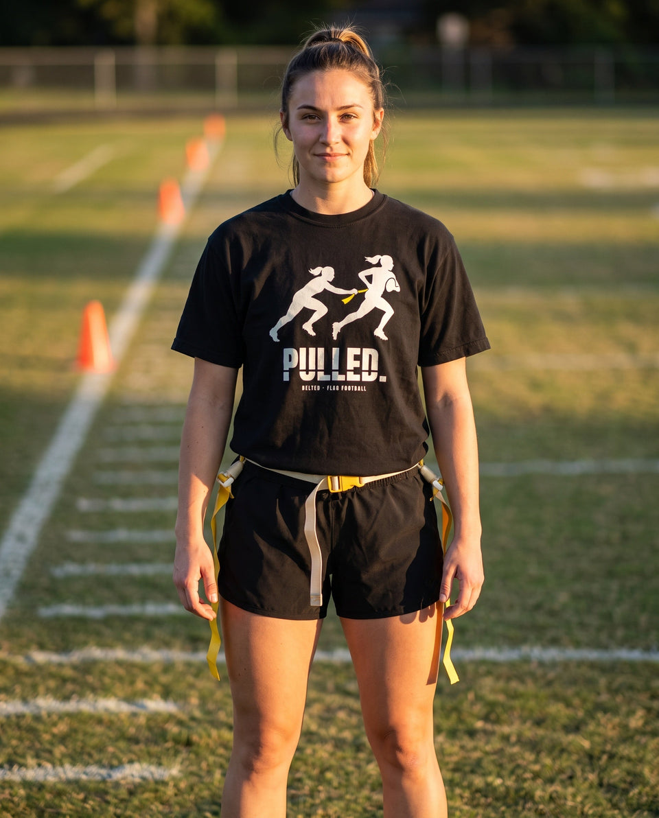 Black Pulled Tee on a model on a field, Ivory two-player pull graphic, PULLED. wordmark, yellow flag, flag belt at waist