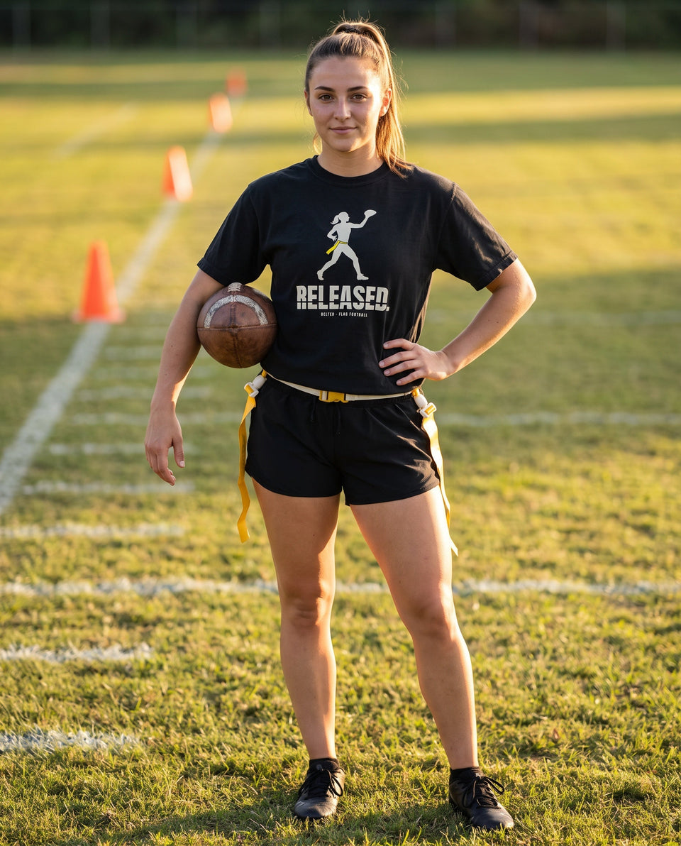 Player in a black Released Tee and black shorts with yellow flag belt, holding a football on a field with cones