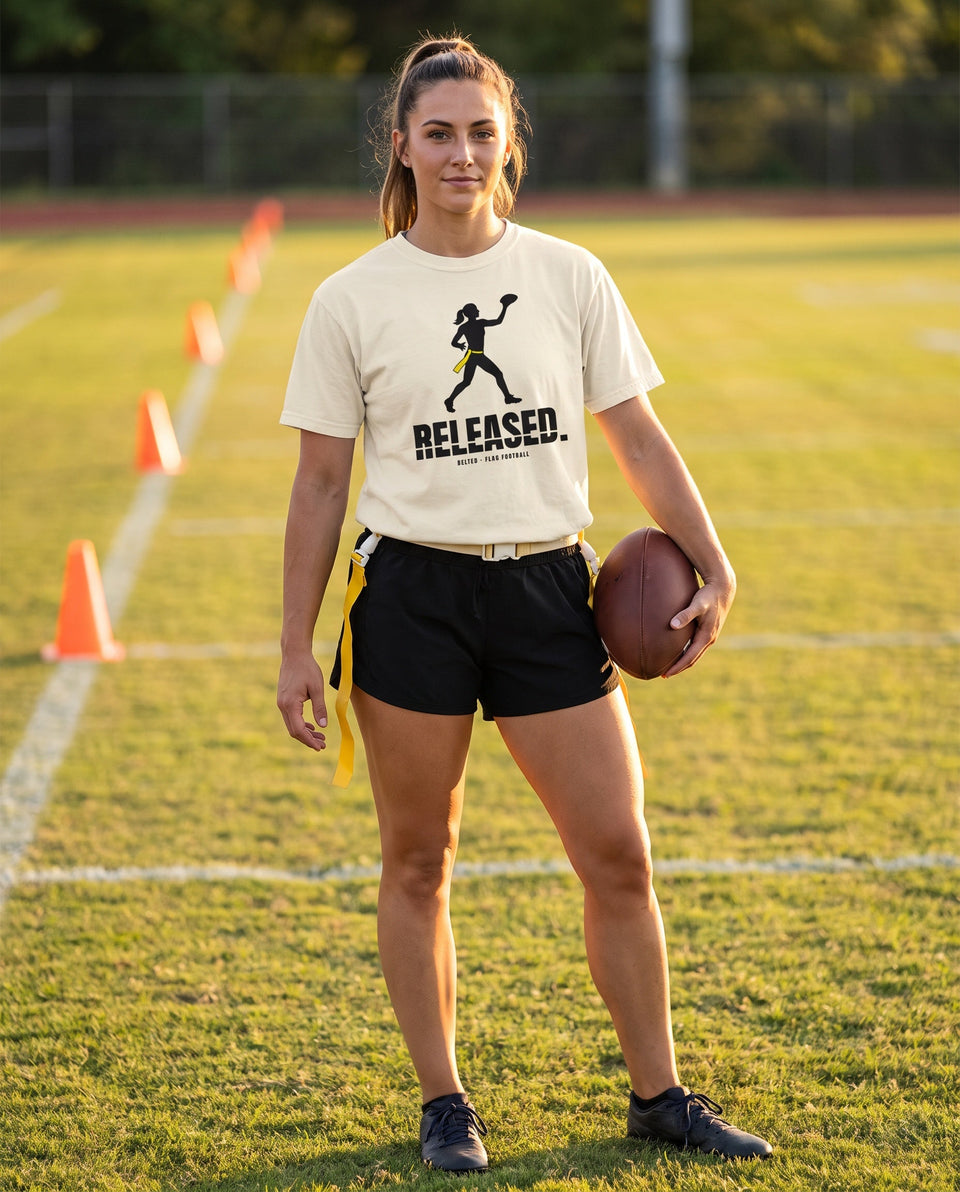 Player in an Ivory Released Tee and black shorts with yellow flag belt, holding a football on a field with cones